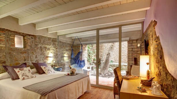 Une chambre avec un mur en pierre, un lit agrémenté de coussins à motifs floraux, un bureau en bois et un fauteuil suspendu.
