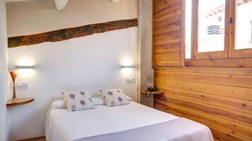 Une chambre à coucher avec une tête de lit en bois, un lit recouvert d’une literie blanche et agrémenté d’oreillers à motifs floraux violets, une table de chevet sur laquelle est posé un vase de fleurs, et une fenêtre donnant sur des immeubles.