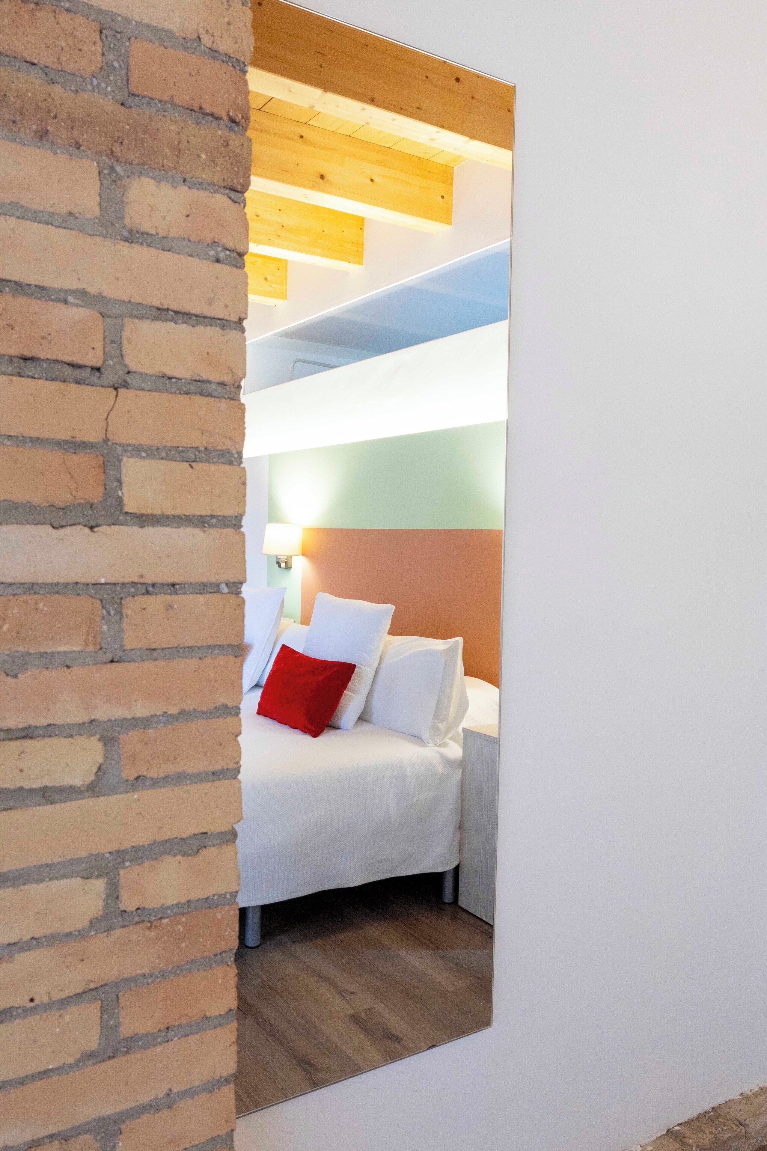 A narrow hallway with a brick wall on the left and a bed with white and red pillows in the room on the right.