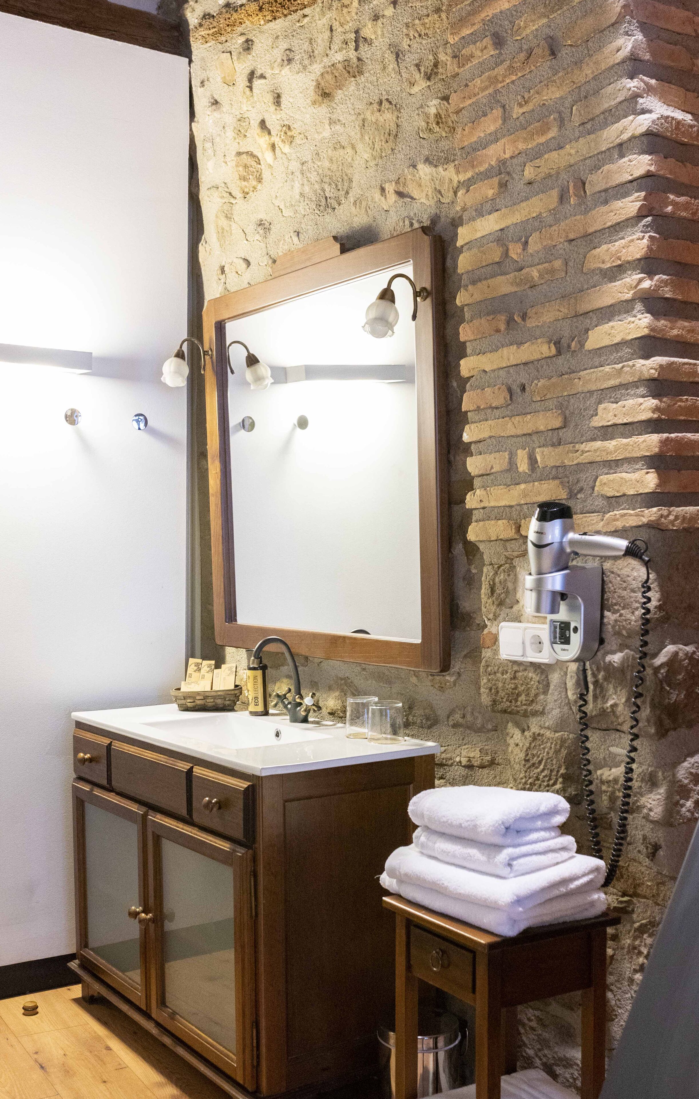 A bathroom with a stone wall, a wooden vanity with a mirror, a hairdryer, and a stack of towels.