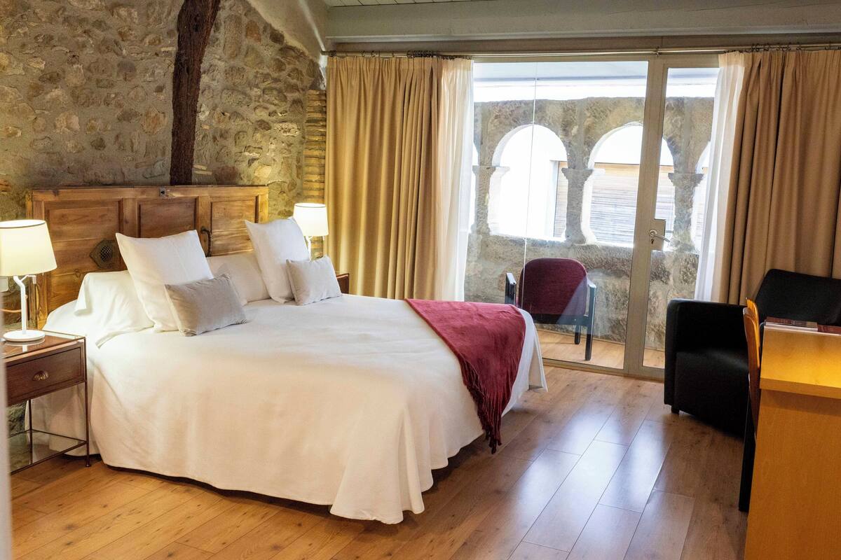 Un dormitorio con una cama grande, cabecera de madera y vistas a una construcción de piedra exterior a través de una puerta de cristal.