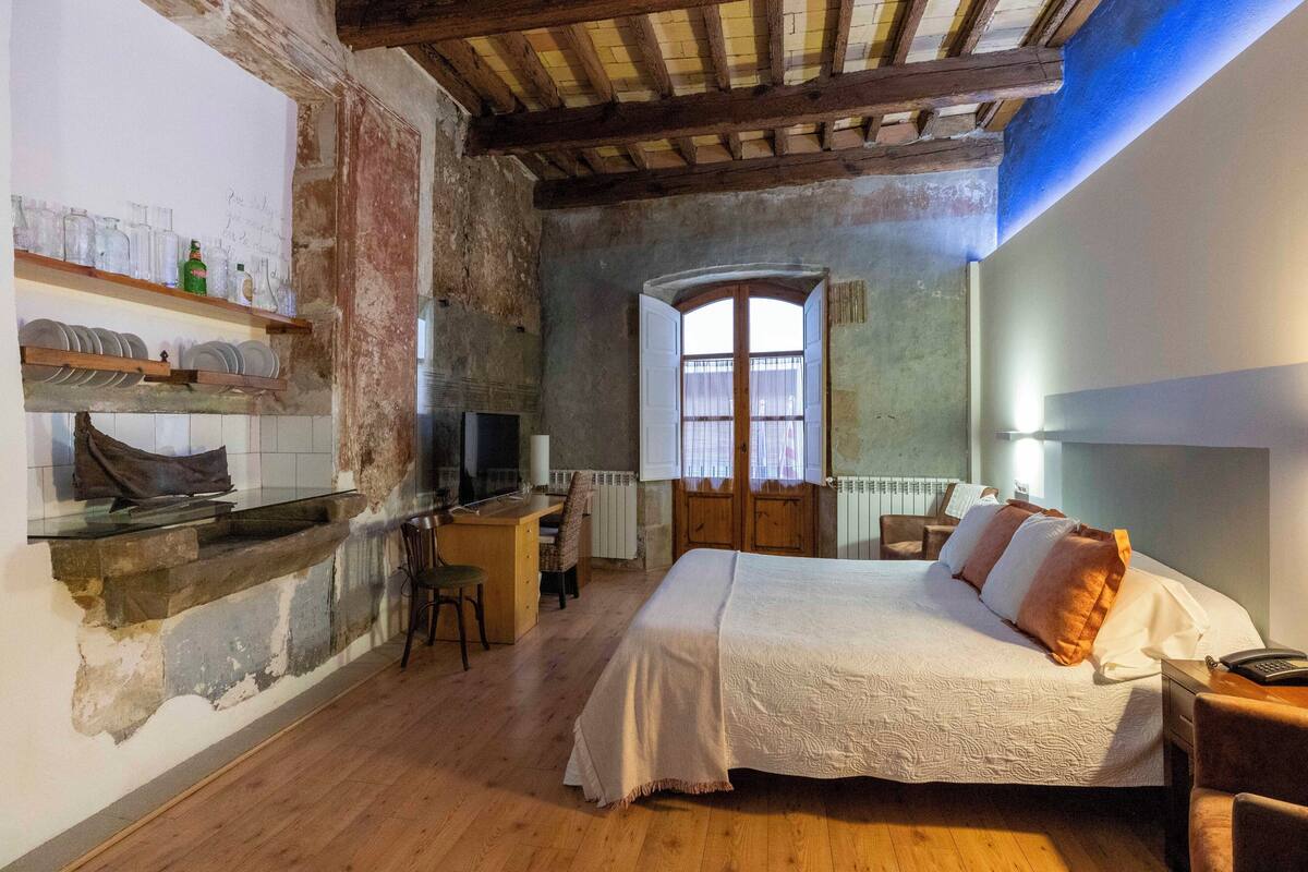 Una habitación rústica con una cama, un escritorio de madera, estantes con vajilla y una ventana.