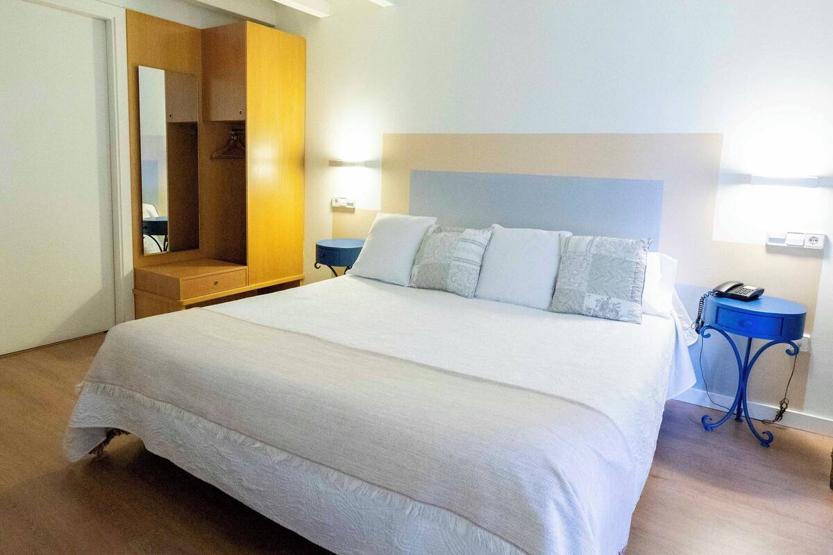 Un dormitorio ordenado con una cama grande, un armario, una mesita de noche con una lámpara y un espejo.