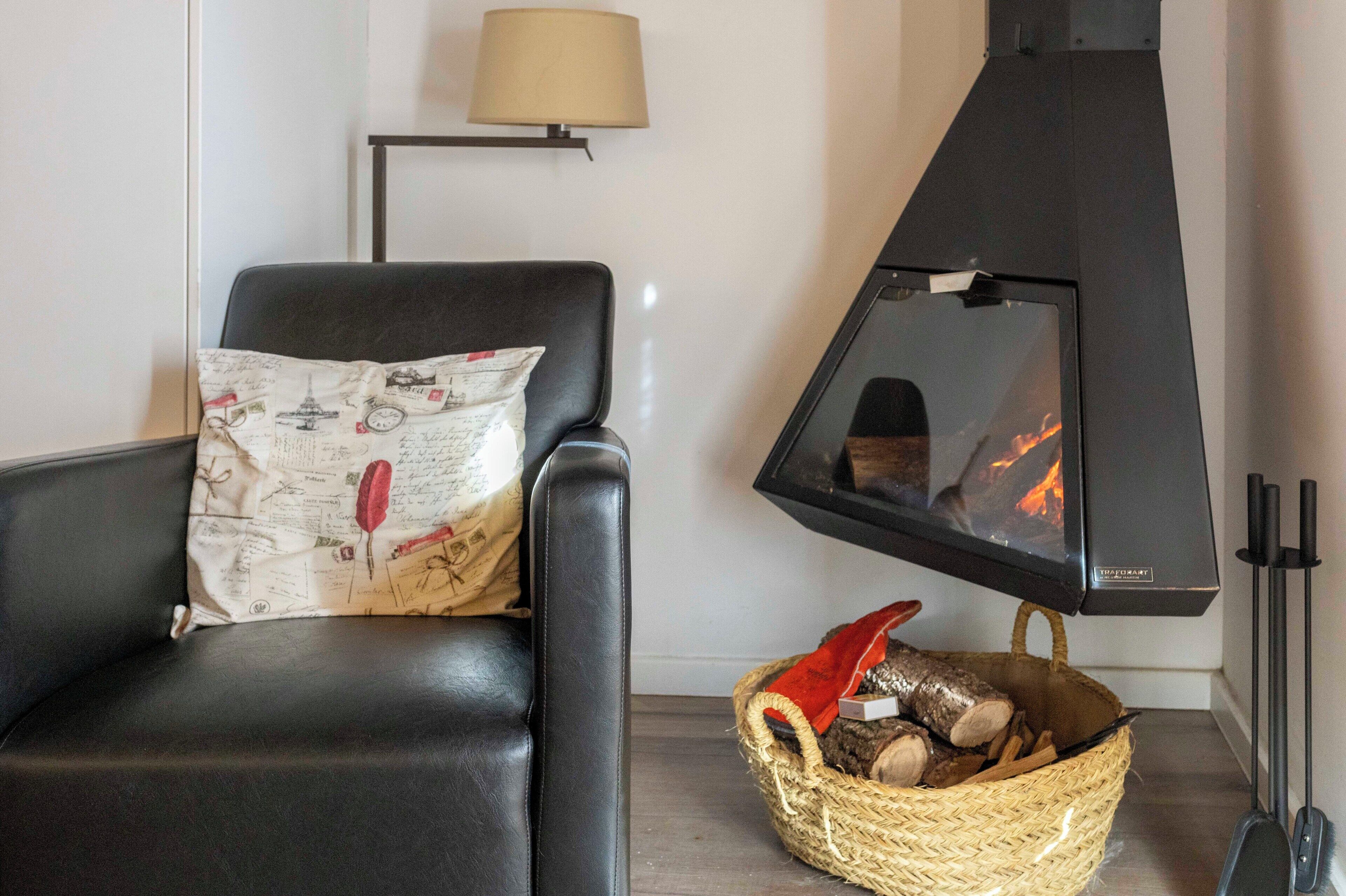 A cozy room with a black leather chair, a woven basket of firewood, and a black wood stove with visible flames.