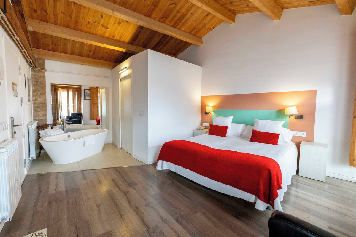 Habitación de hotel con una cama grande, baño independiente con bañera, vigas de madera en el techo y vistas a la ciudad.