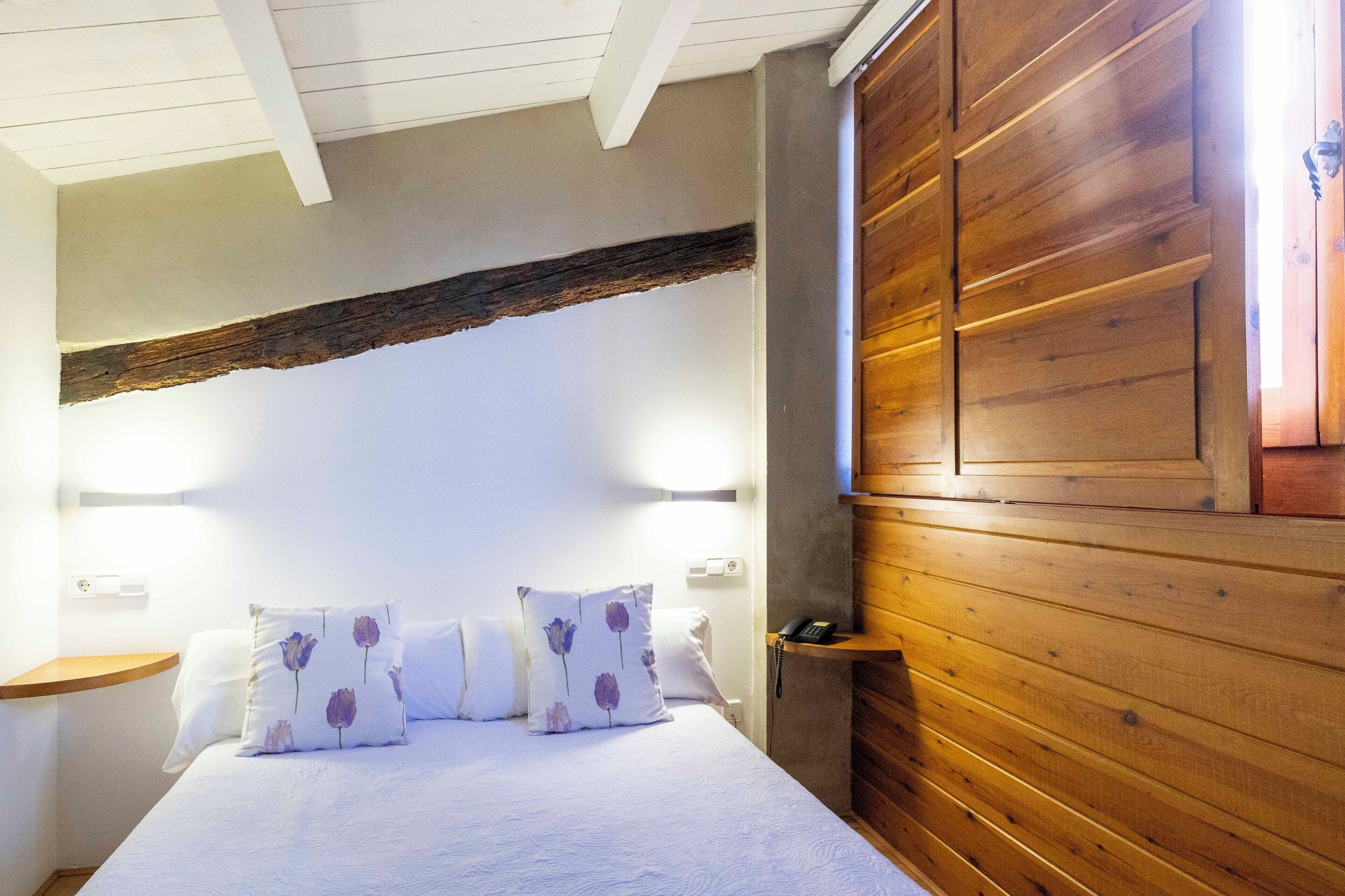 Un dormitorio con cabecera de madera, cama con ropa de cama blanca y almohadas estampadas con flores moradas, una mesita de noche y una mesilla.
