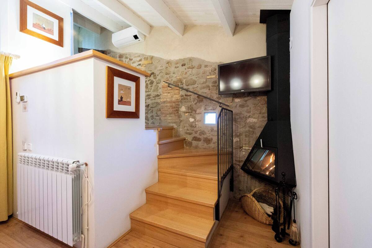 Una escalera de madera conduce a una plataforma, con un muro de piedra, una chimenea y un televisor fijado a la pared.