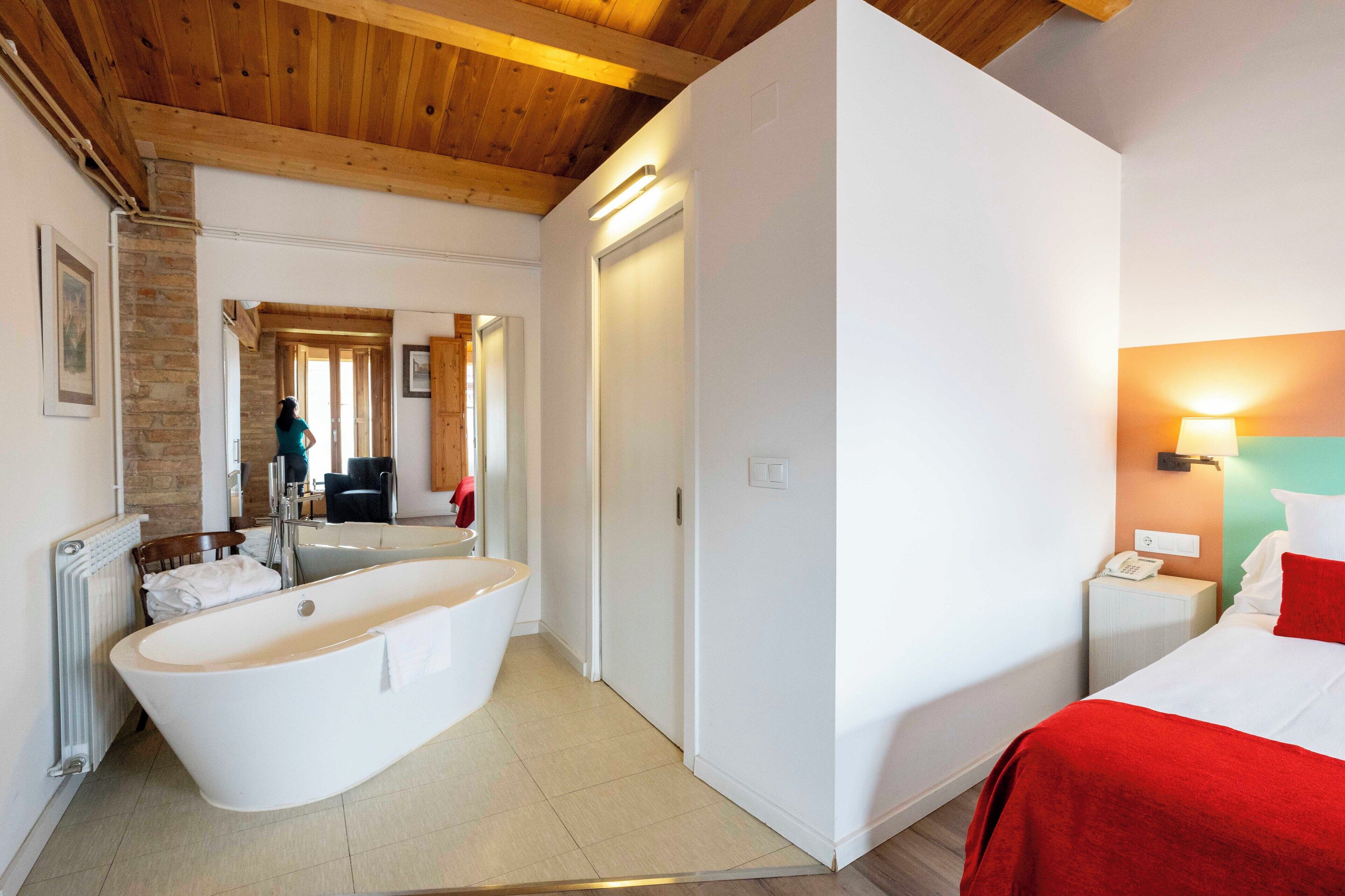 A modern hotel room with a large bathtub, a bed with red bedding, and a wooden ceiling.