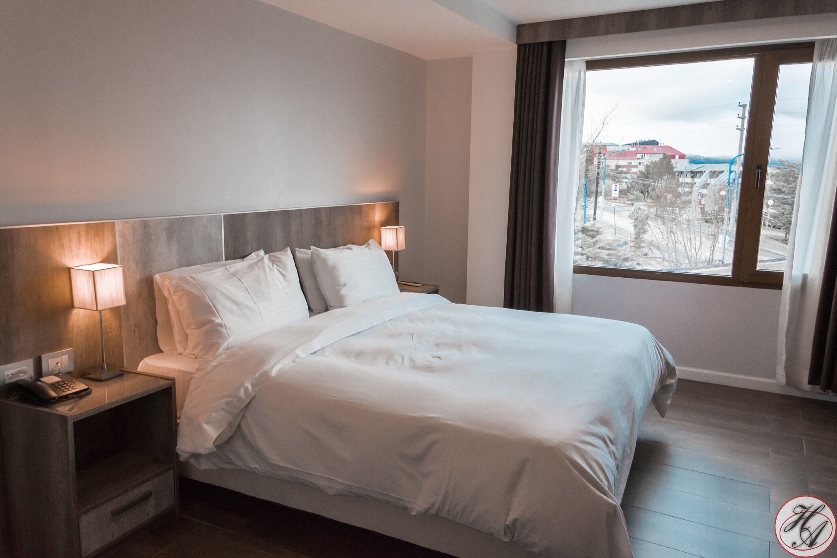 Ein Hotelzimmer mit einem großen Bett, zwei Nachttischlampen, einem Nachttisch mit Telefon und einem Fenster mit Blick auf Gebäude und Bäume.