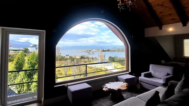 A cozy living room with a large arched window offering a scenic view of a lake and mountains. -  Hostería y Restaurante América (Ushuaia)