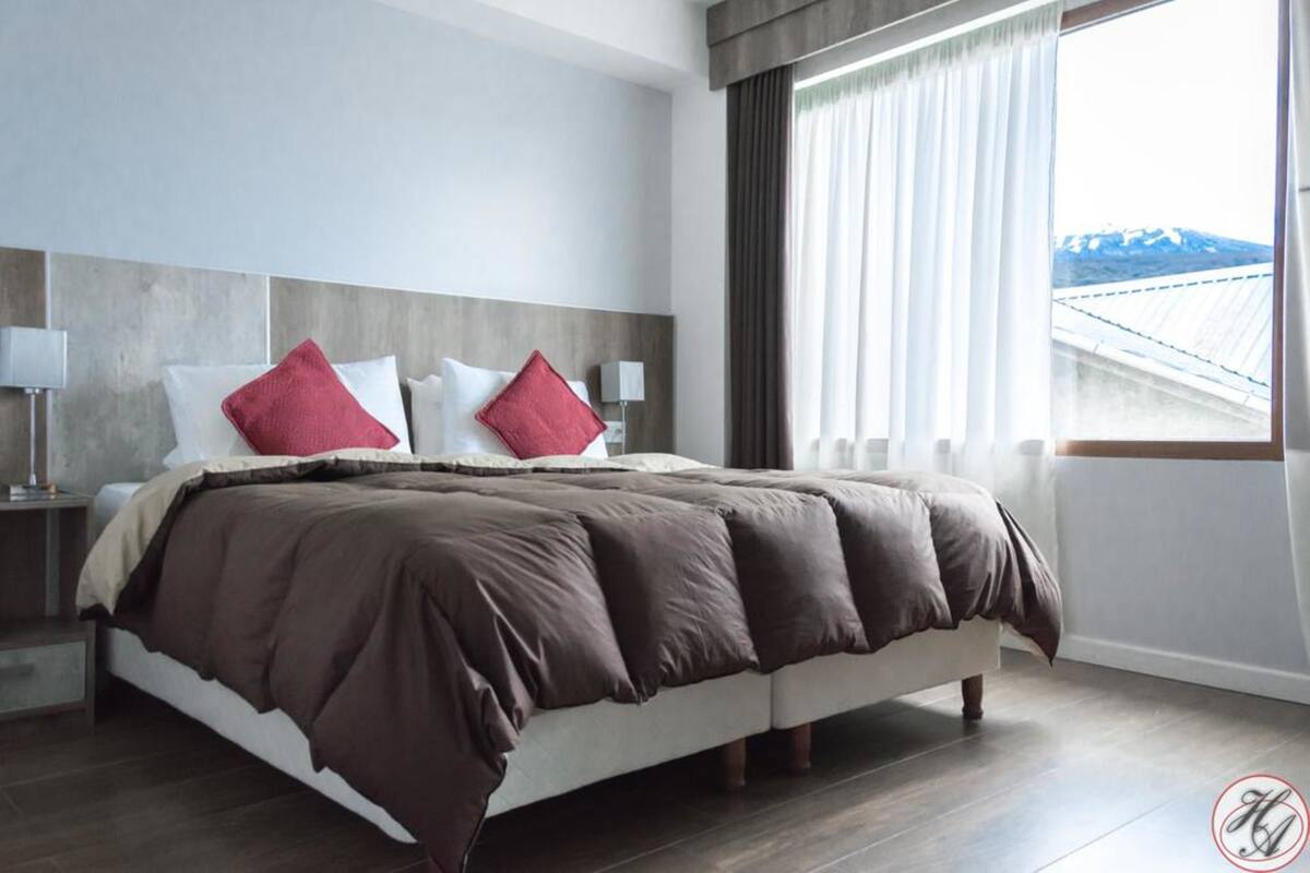 Habitación de hotel moderna con una cama grande, dos mesitas de noche y vistas a una montaña desde la ventana.