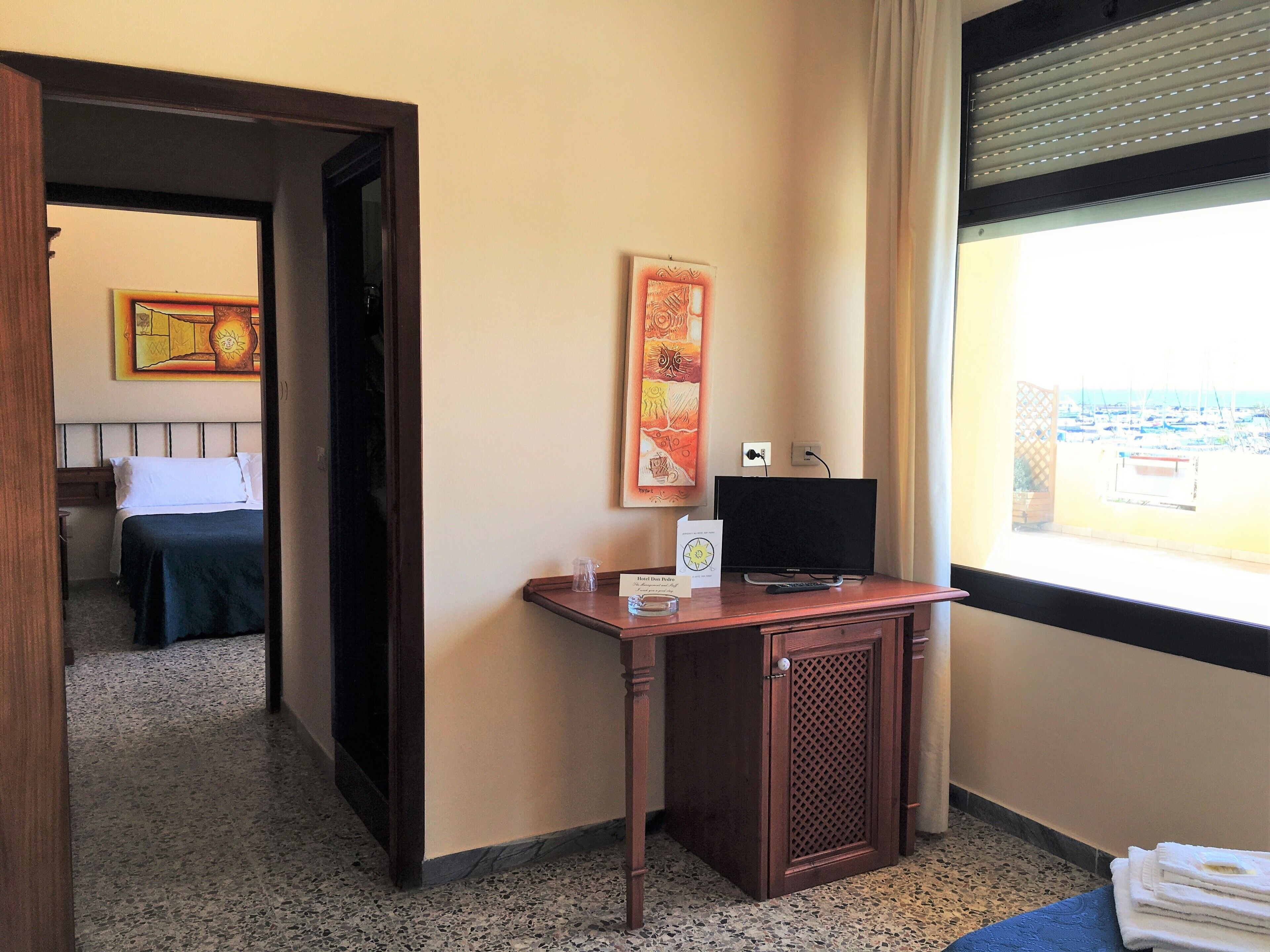 A hotel room with a bed, a desk with a television, a window with a view of boats, and a painting on the wall.
