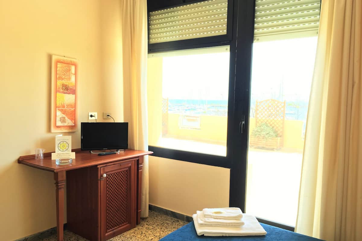 A hotel room with a wooden desk, a television, a window with a view of the sea, and a bed with a blue bedspread.