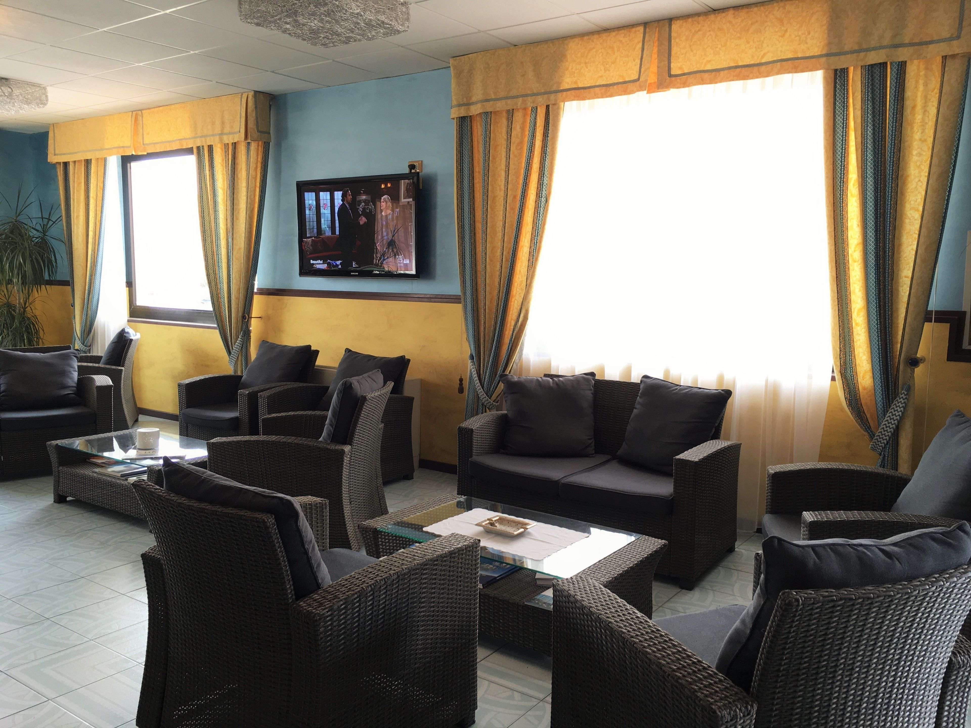 A hotel lobby with wicker furniture, a flat-screen TV, and large windows with curtains.