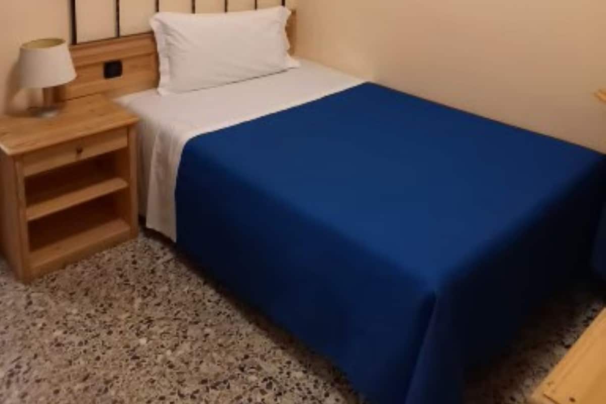 A single bed with a blue bedspread, a wooden nightstand with a lamp, and two framed pictures on the wall.