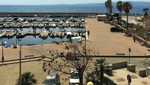 Un puerto deportivo con numerosas embarcaciones amarradas, un aparcamiento con coches y vistas al mar y colinas lejanas.