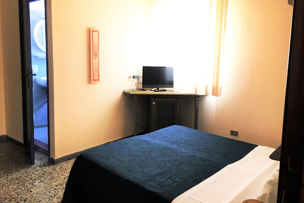 A hotel room with a bed, a desk with a computer, a door leading to a bathroom, and a window with curtains.