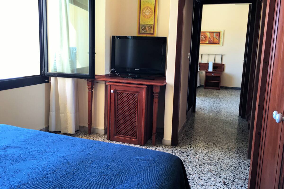 A hotel room with a bed, a television on a wooden stand, a window with curtains, and a hallway leading to another room.
