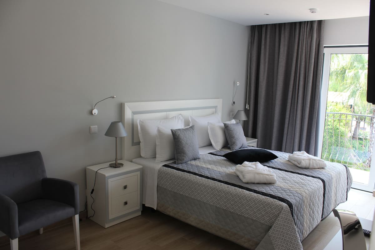 Moderni hotellihuone, jossa on siististi pedattu sänky, yöpöytä lampun kanssa, tuoli ja näkymä vihreälle alueelle ikkunasta.