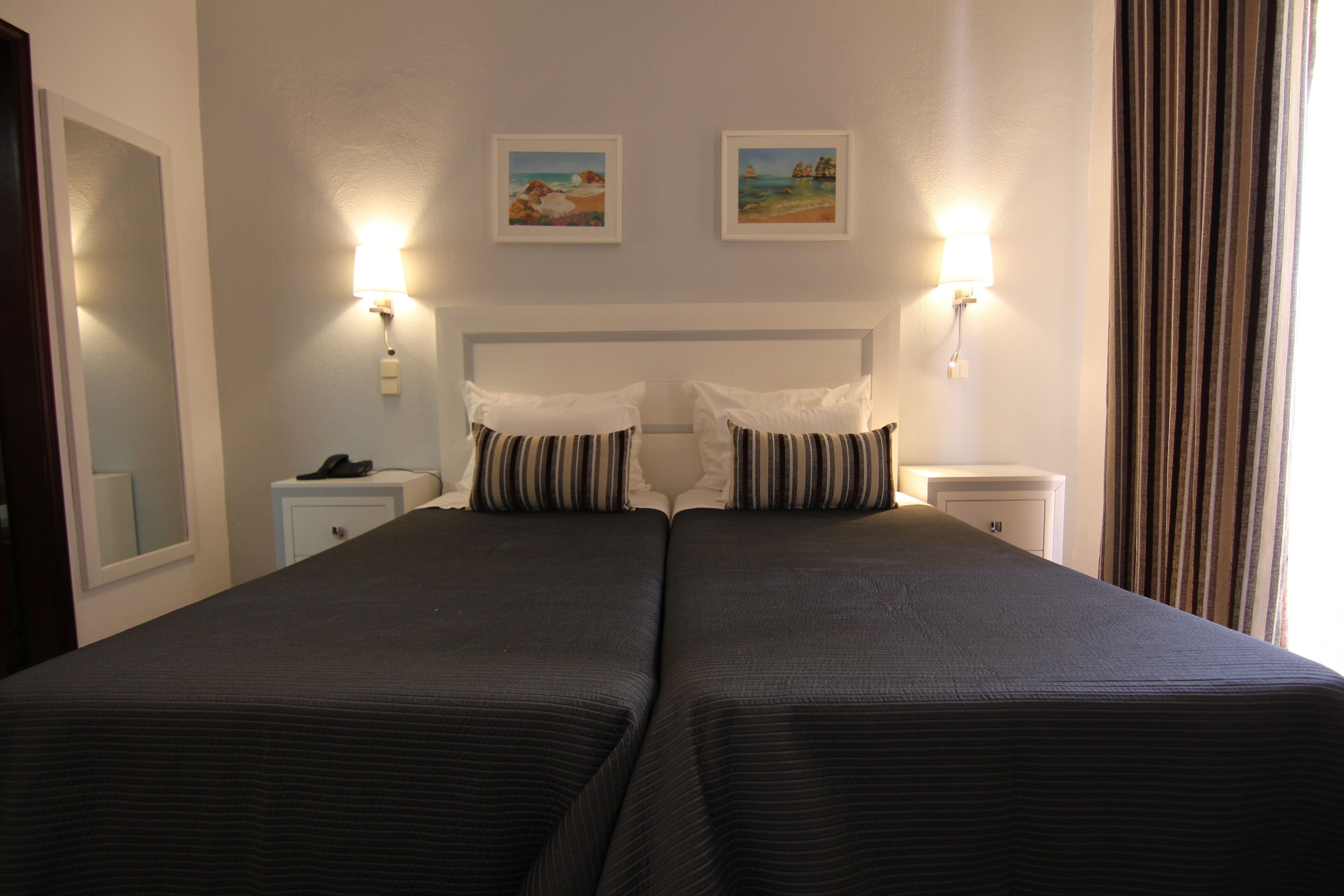 Een tweepersoonsbed met gestreepte beddengoed, twee nachtkastjes, een wandlamp en twee ingelijste foto's aan de muur.