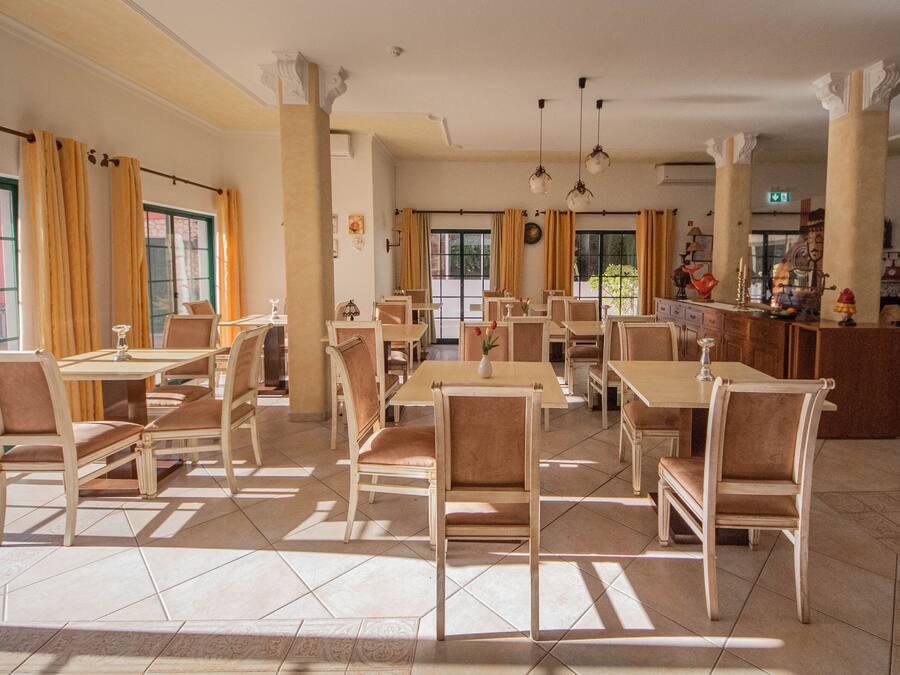 A spacious dining area with multiple tables and chairs, large windows with curtains, and a bar area with various items.
