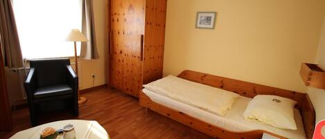 A small room with a wooden bed, a chair, a table with a cupcake and a glass, and a wardrobe.