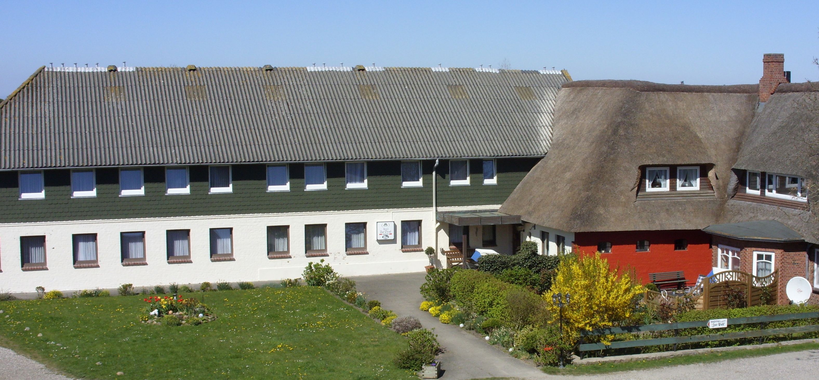 A building with a green roof and white walls, a thatched-roof structure, and a garden with flowers and shrubs.