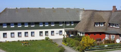 A building with a green roof and white walls, a thatched-roof structure, and a garden with flowers and shrubs.