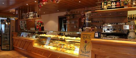 A bakery display case with various baked goods, a bar area with shelves of bottles, and a wooden counter with a sign reading "BAR Nizza".