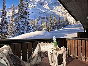 A wooden deck with a rustic table and chairs, overlooking a snowy mountain landscape with evergreen trees. - Nigritella (Selva di Cadore)