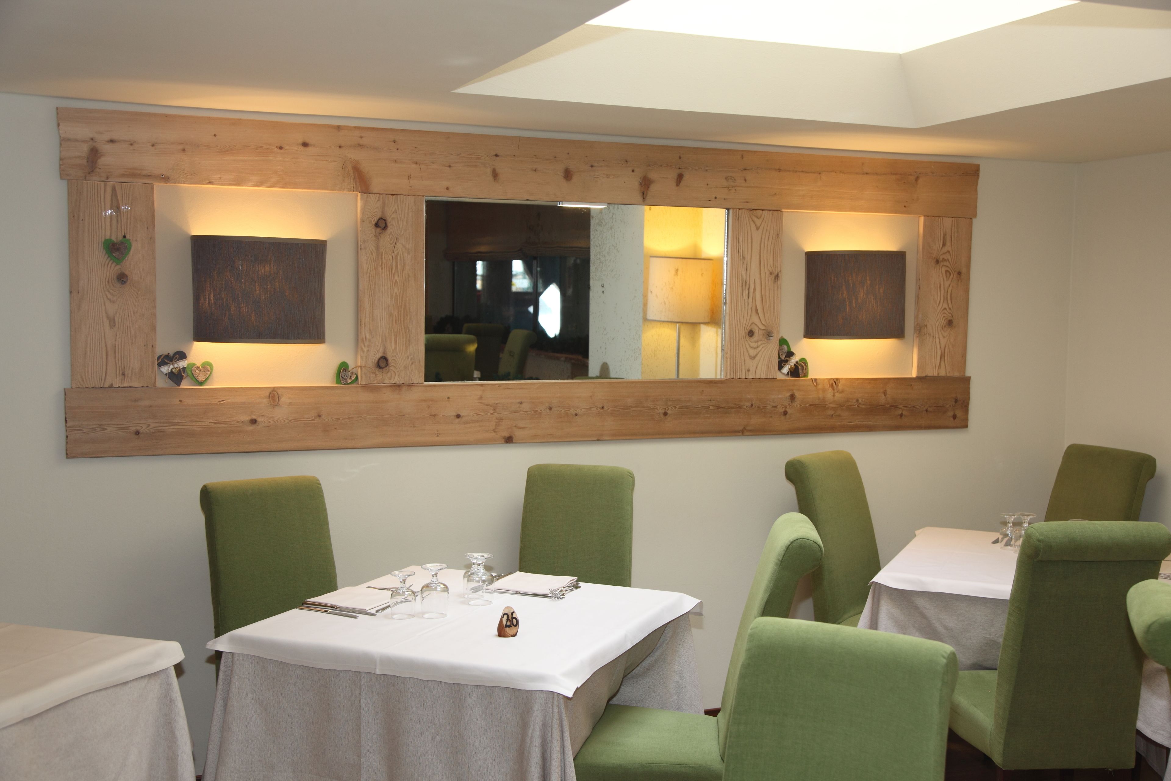 A dining area with a wooden-framed mirror, green upholstered chairs, and a table set with a white tablecloth, wine glasses, and cutlery.