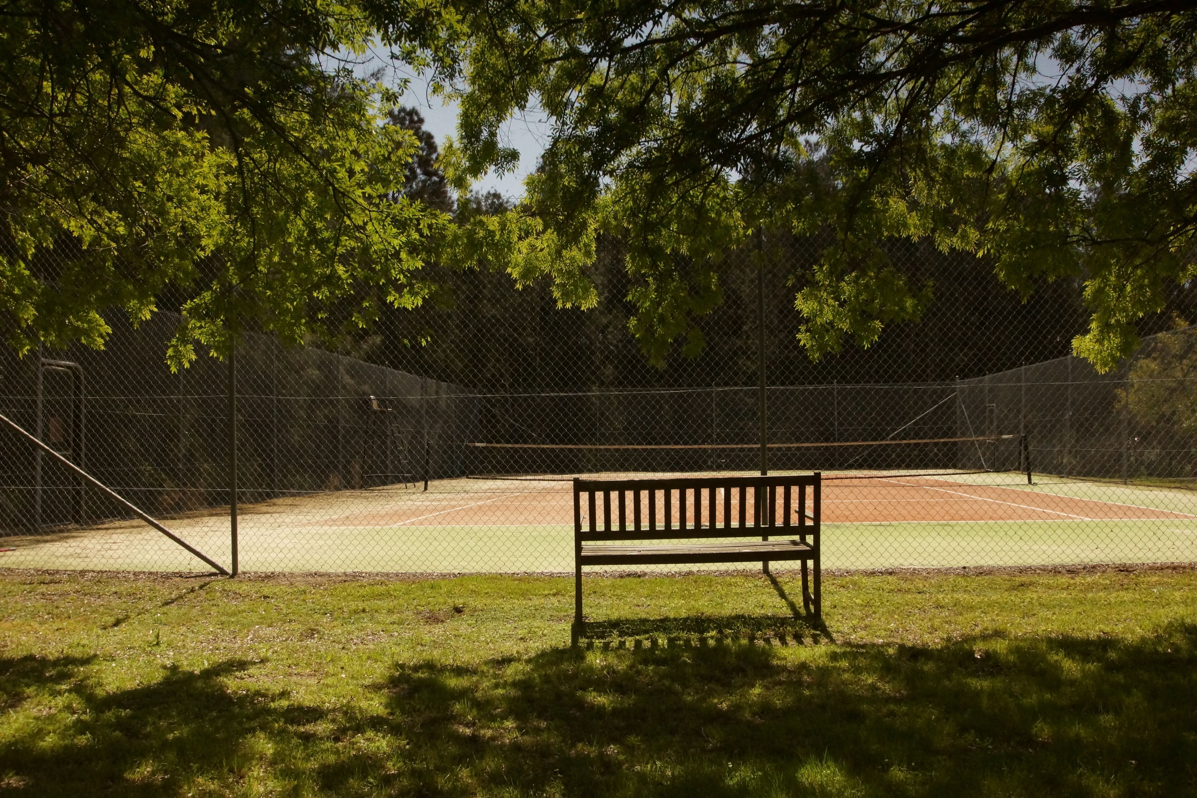 A tennis court with a bench in the foreground, surrounded by a chain-link fence and trees.