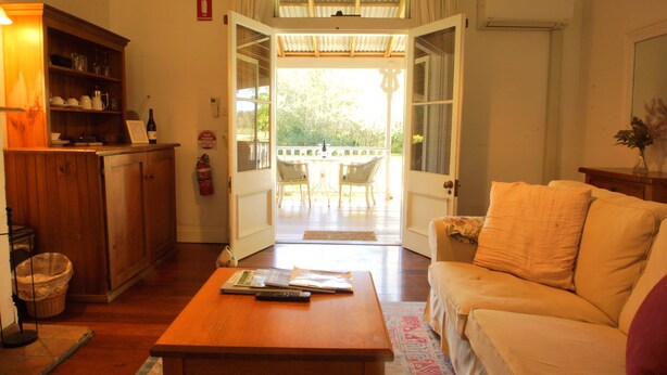 An open door leading to a balcony with a table and chairs, a wooden cabinet with dishes, and a sofa in the living area. - The Carriages (Pokolbin)
