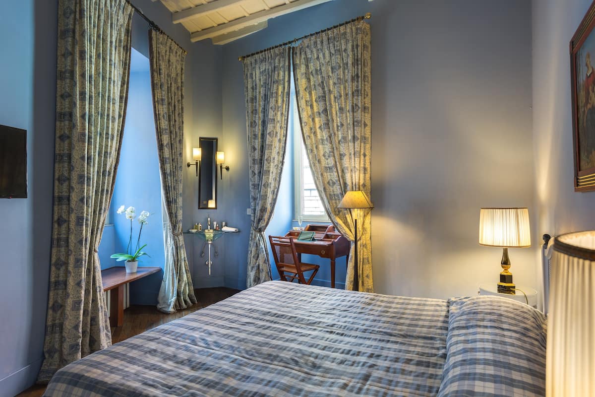 Ein Schlafzimmer mit einem Bett, einem Schreibtisch und einem Stuhl. Vor den Fenstern hängen Vorhänge, auf dem Schreibtisch steht eine Lampe und am Nachttisch ebenfalls eine Lampe.