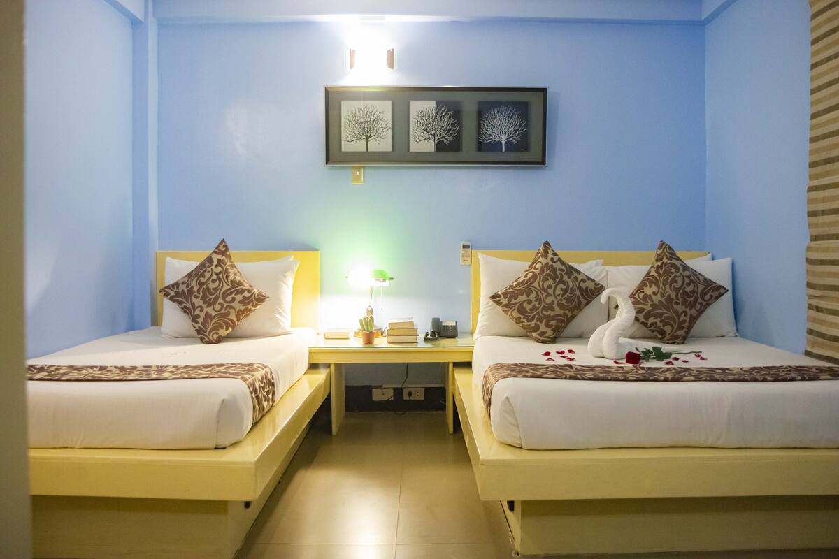 A hotel room with two beds, each with patterned pillows and a swan decoration. A nightstand with a lamp and books is between the beds.