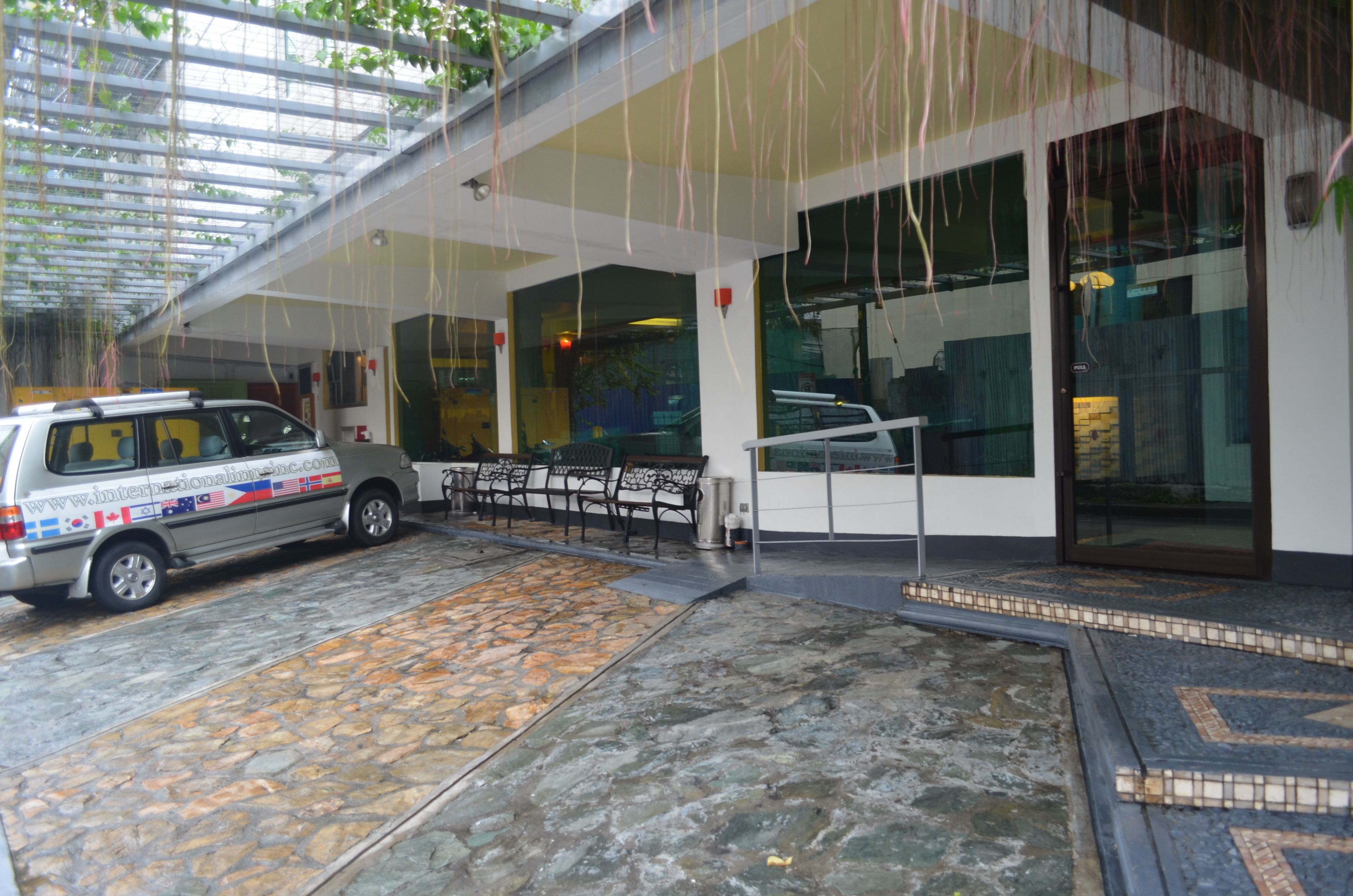 A covered outdoor area with a stone floor, a parked van, and a building entrance with glass doors and windows.