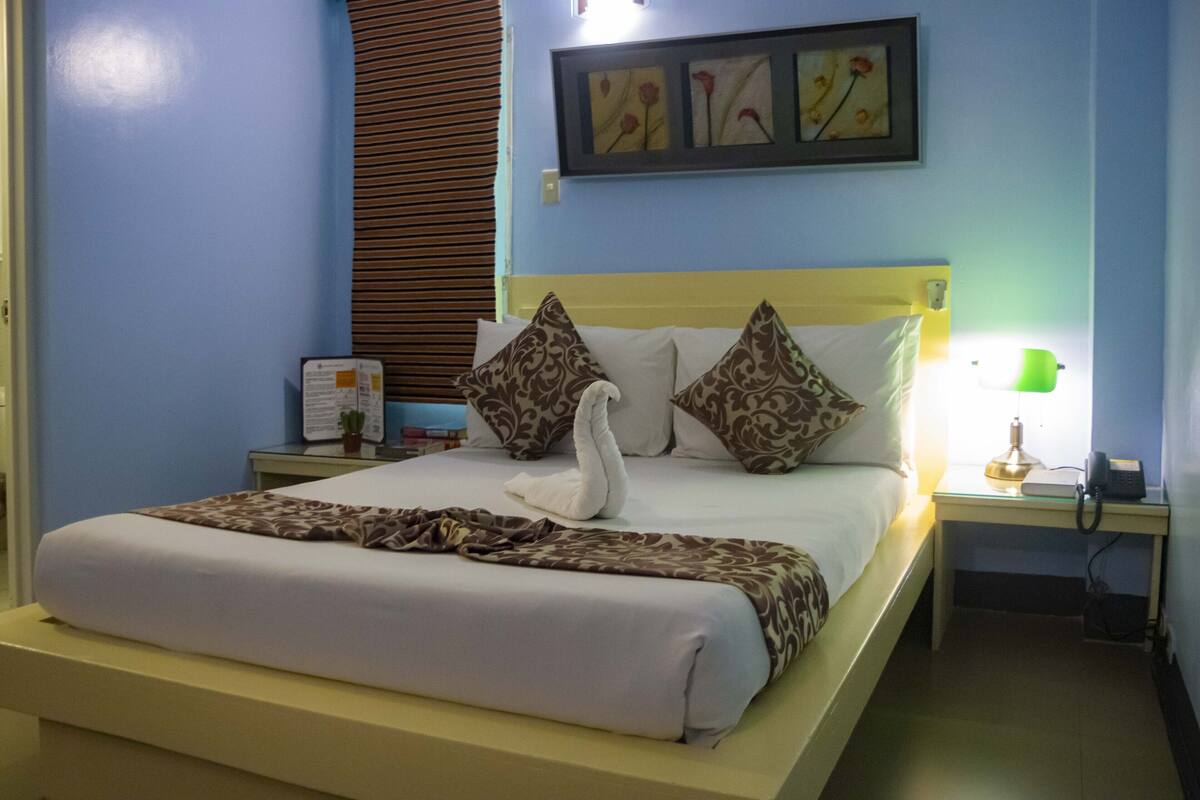 A neatly made bed with a swan-shaped pillow, a bedside lamp, a framed picture on the wall, and a small table with a phone and a plant.