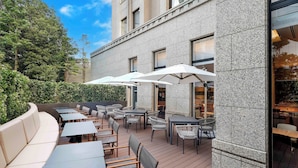 An outdoor patio with tables and chairs, a curved white sofa, and a building with large windows and a stone facade. - Hotel Trusty Tokyo Bayside (Tokyo)