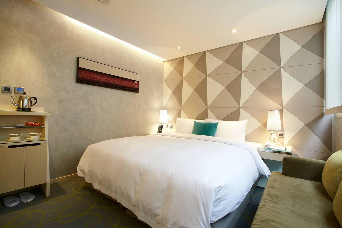 Hotelkamer met een groot bed, een nachtkastje met een lamp, een klein bureau met een waterkoker, en een muur met geometrische patronen.