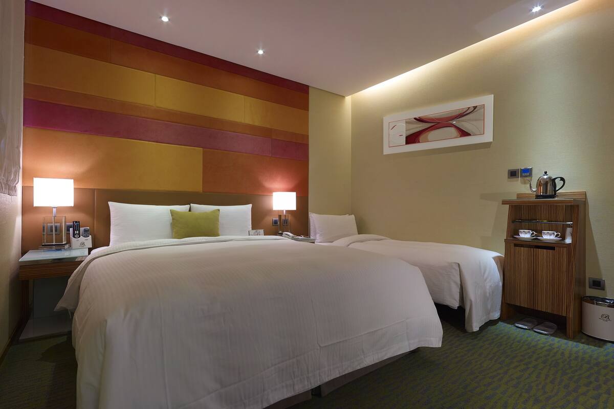 Een hotelkamer met een groot bed, nachtkastjes, een lamp, een klein kastje met kopjes en een kunstwerk aan de muur.
