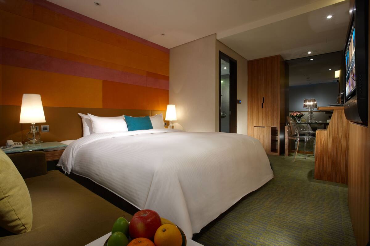 Een hotelkamer met een groot bed, twee bedlampen, een bureau met een lamp en een fruitschaal op de bijzettafel.