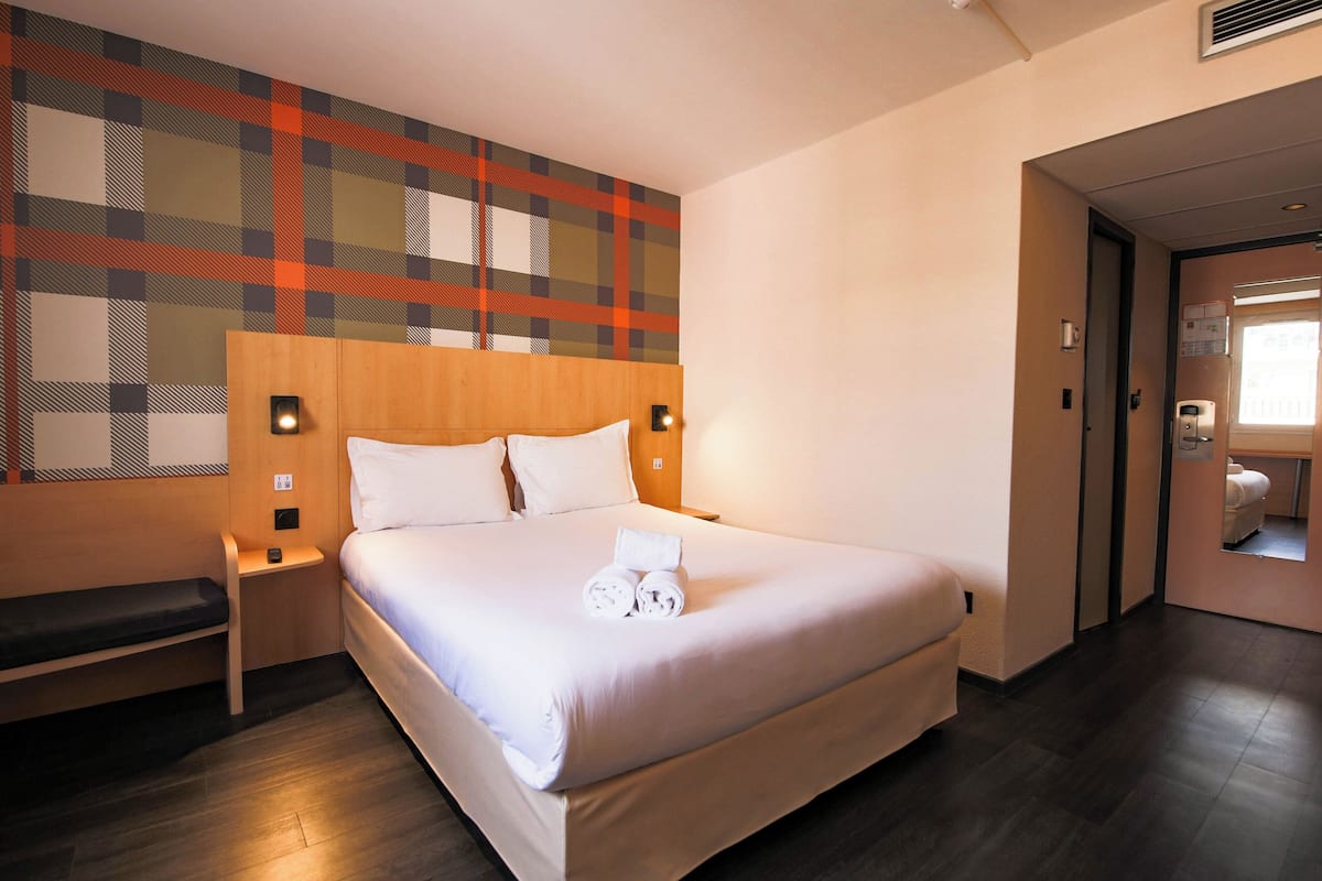 Una cama impecable con sábanas blancas y toallas enrolladas, cabecera de madera y pared con estampado escocés.