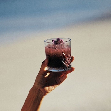 A hand holding a glass with a drink, set against a beach background.
