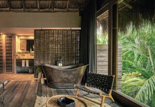 A bathroom with a freestanding bathtub, a wooden chair, and a large window with a view of tropical foliage.
