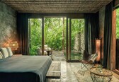 A modern bedroom with a large bed, a seating area, and a view of lush greenery through large windows.