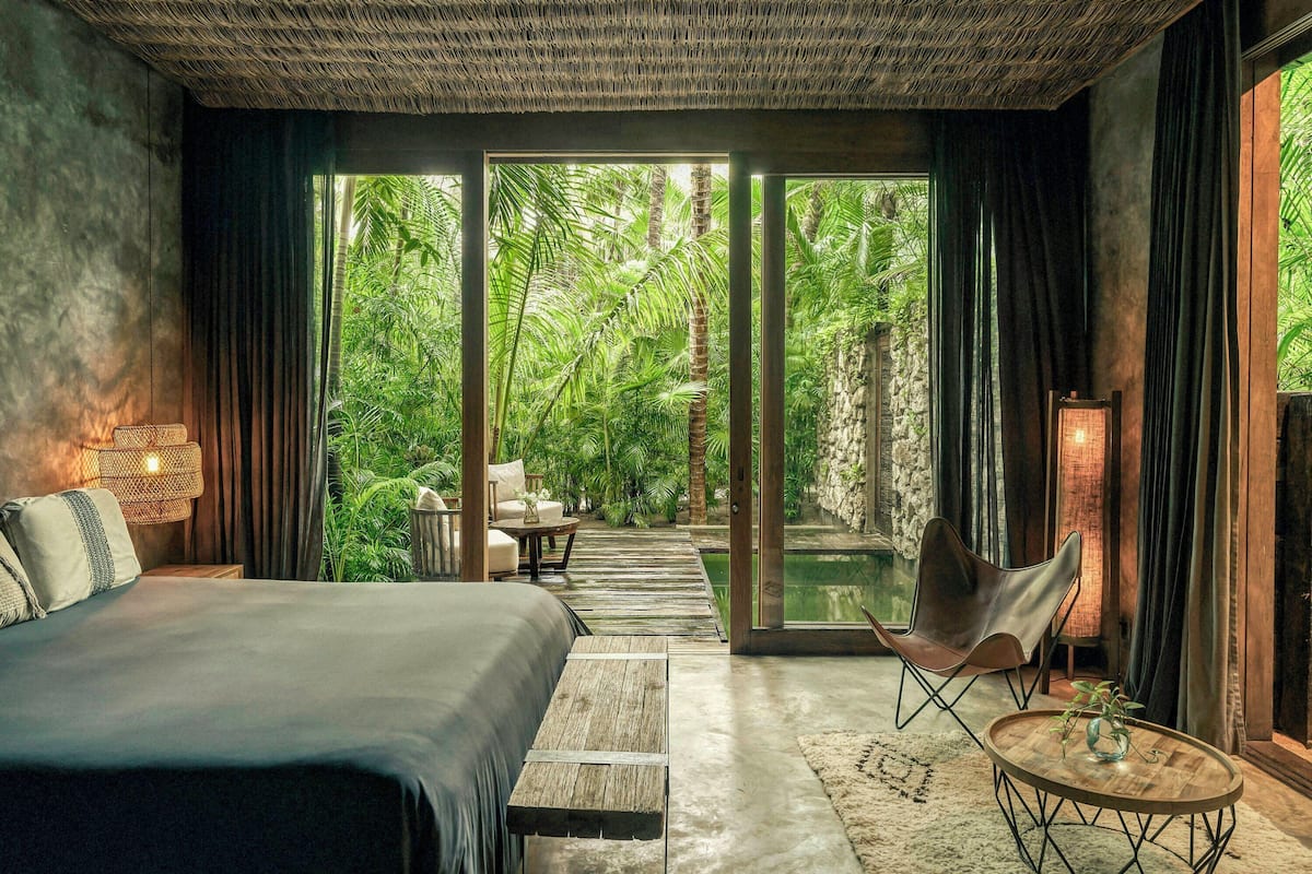 A modern bedroom with a large bed, a seating area, and a view of lush greenery through large windows.