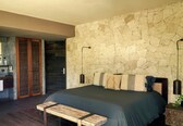 A bedroom with a stone wall, a bed, a bench, a nightstand, and a mirror.