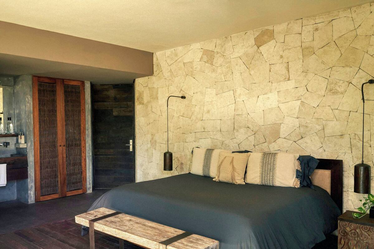 A bedroom with a stone wall, a bed, a bench, a nightstand, and a mirror.