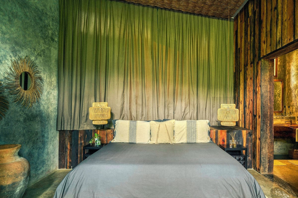 A room with a bed, green curtains, wooden walls, and two wall-mounted lamps.