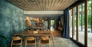 A dining area with a wooden table and chairs, a kitchen in the background, and large windows with curtains.